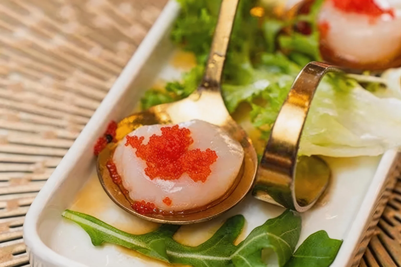 Scallops with tobiko served in golden spoons, on a white plate with salad.