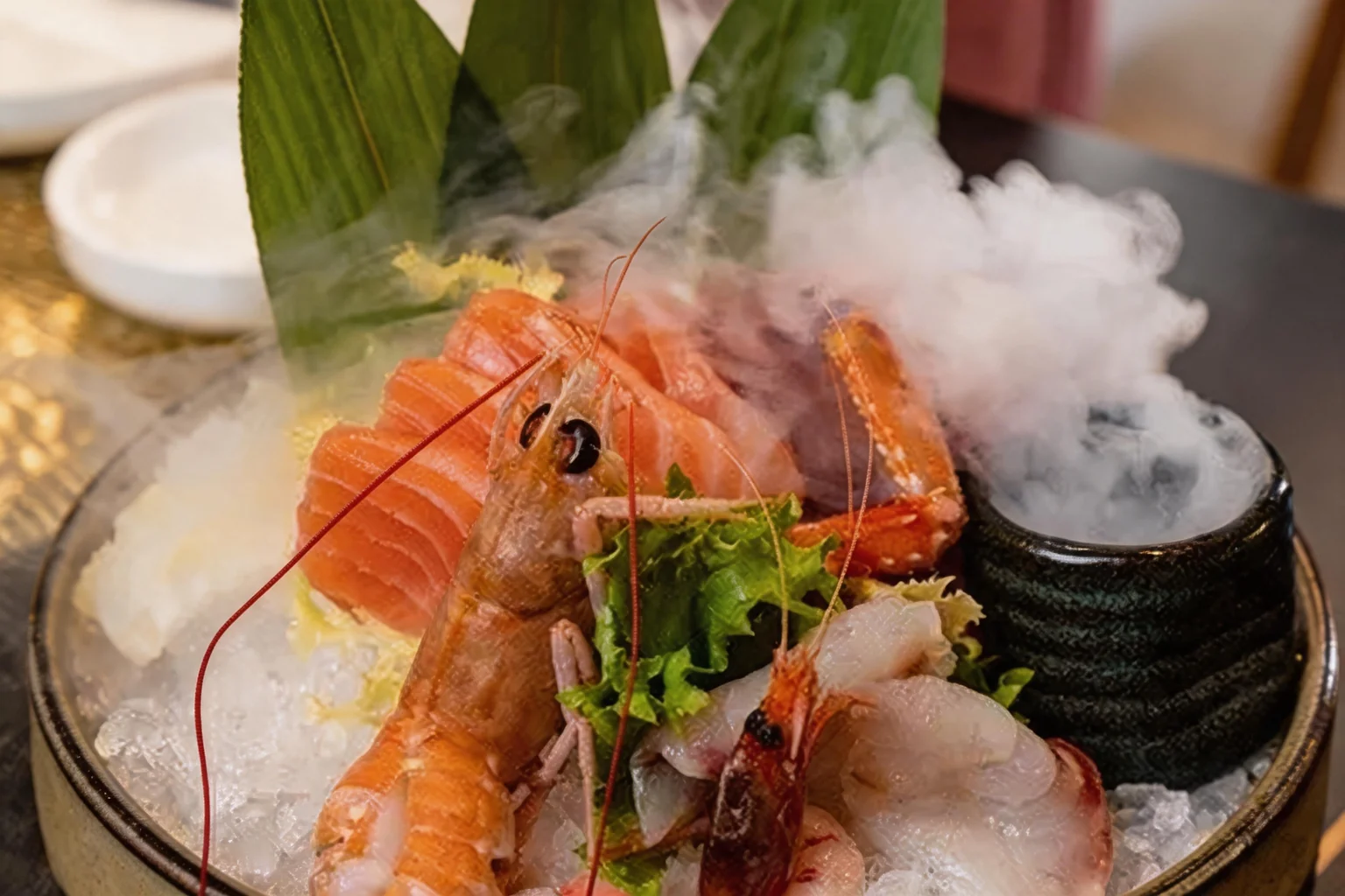 Salmon and white fish sashimi with red prawns and langoustines on ice, with green leaves and dry-ice mist.